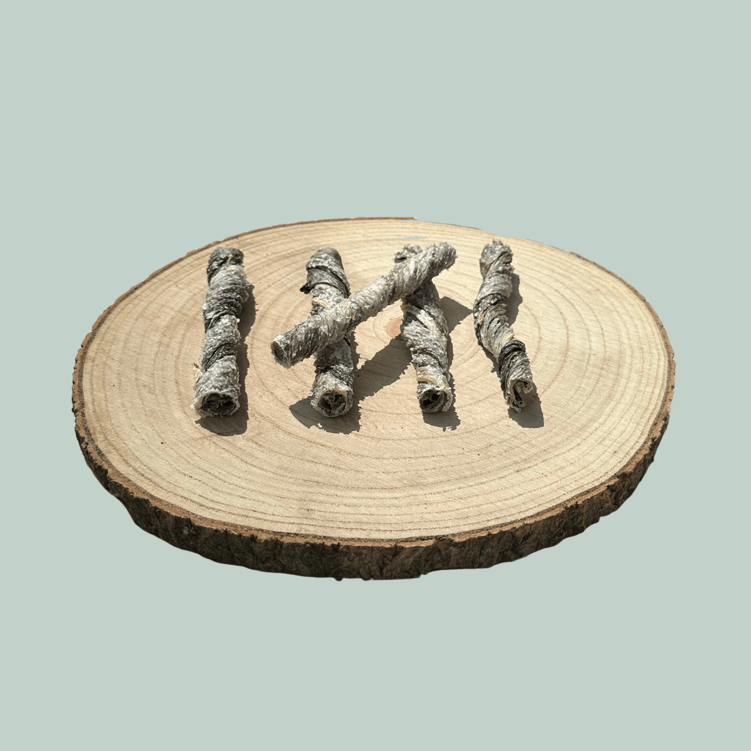 Side view of 5 dried salmon skin dog treats on a natural wooden slice plate against a jade green background