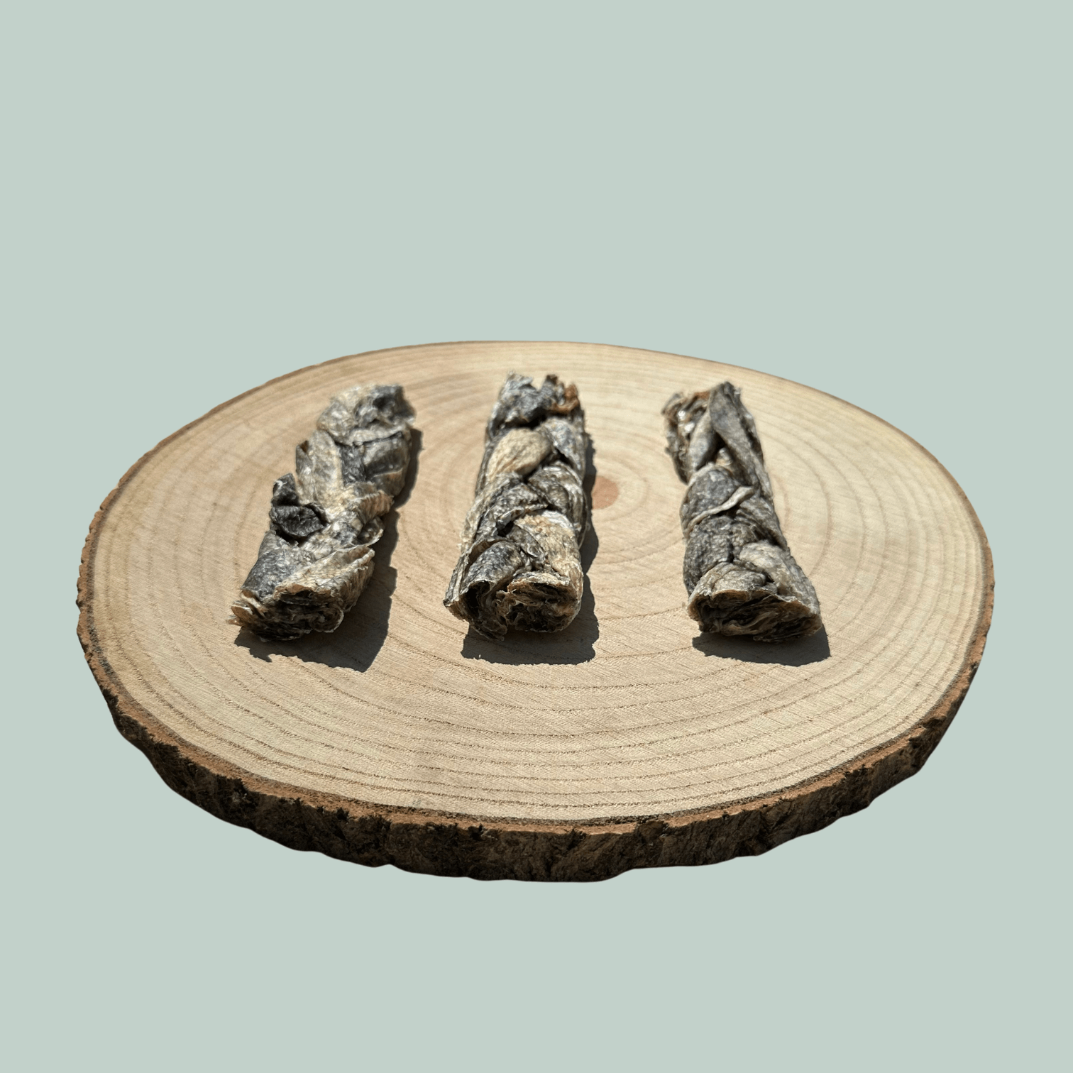 Side view of 3 natural cod skin braid dog chews on a wooden slice plate against a jade green background