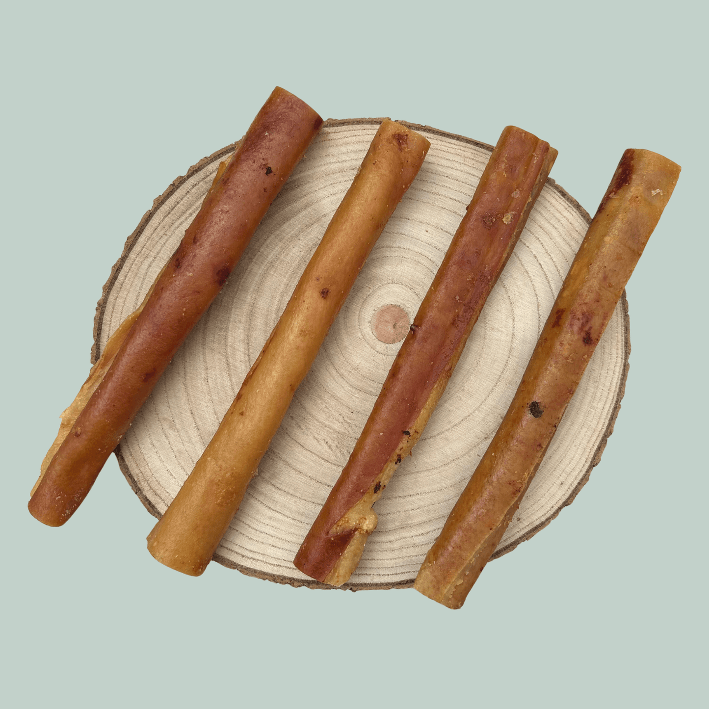 Four pork rind roll natural dog treats on a wooden slice plate against a jade green background.