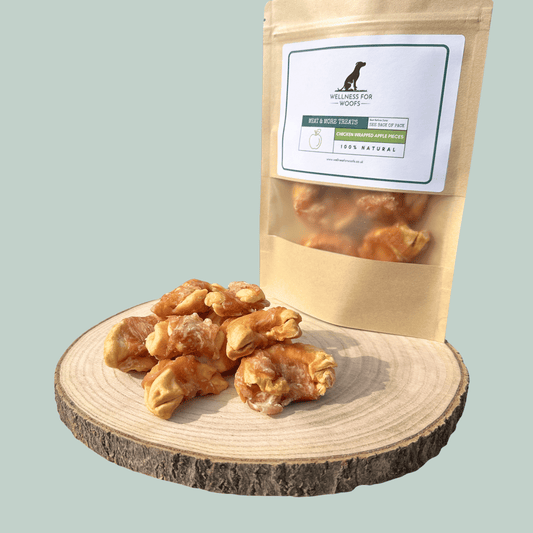 Slices of apple wrapped in dried chicken in front of a natural dog treat pouch on a wooden slice on a green background