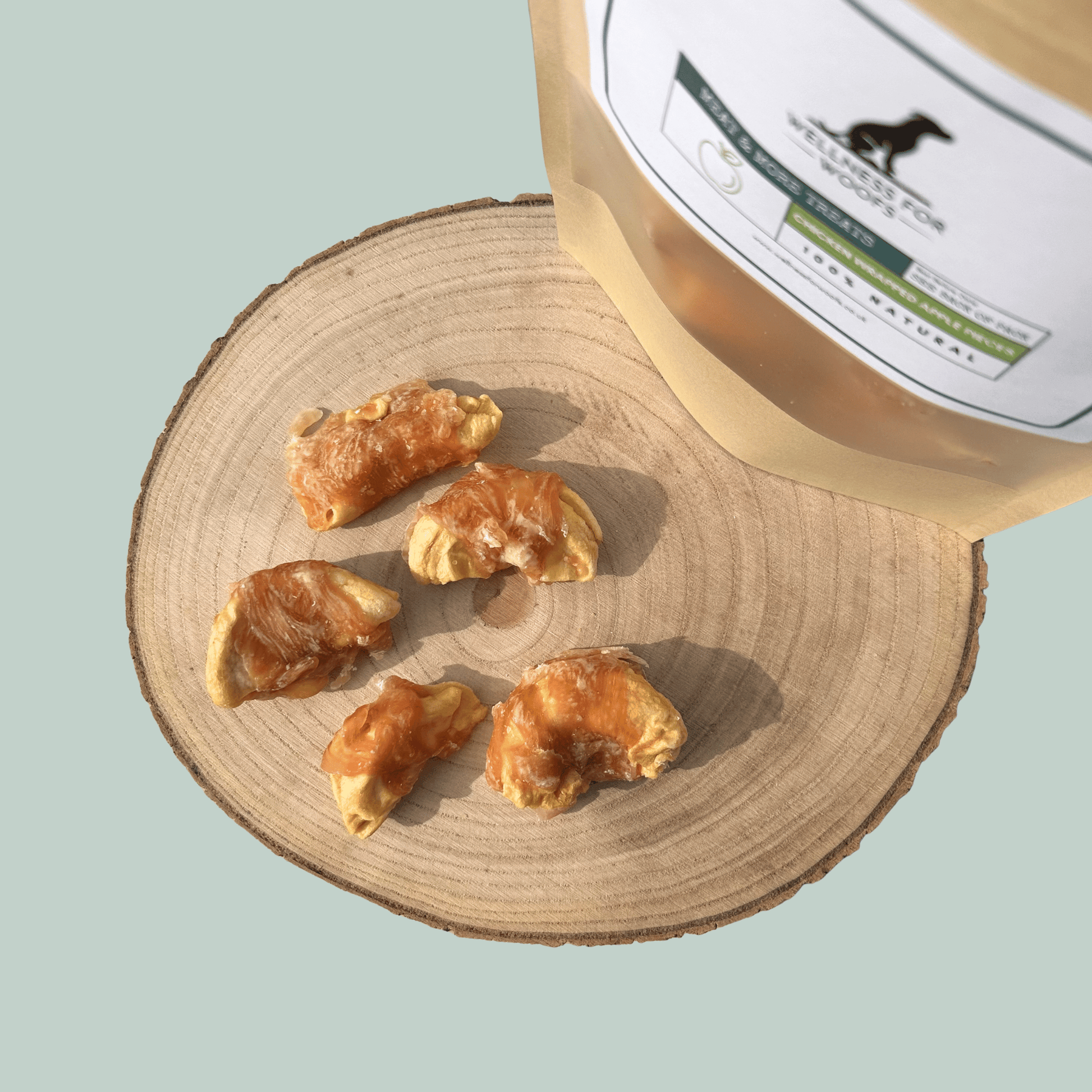 Slices of apple wrapped in dried chicken in front of a natural dog treat pouch on a wooden slice on a green background