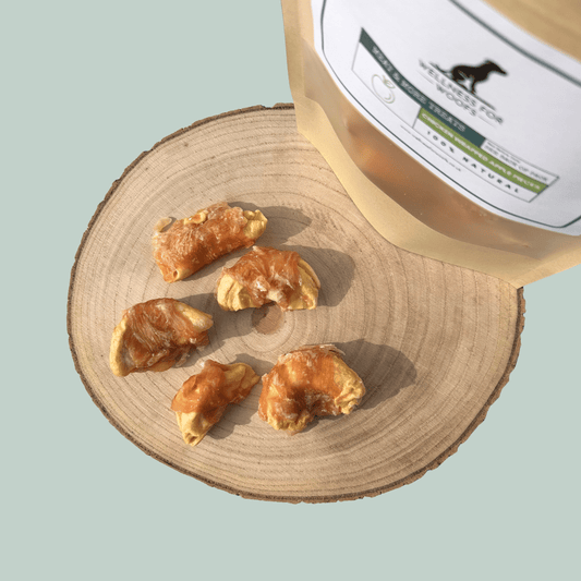 Slices of apple wrapped in dried chicken in front of a natural dog treat pouch on a wooden slice on a green background