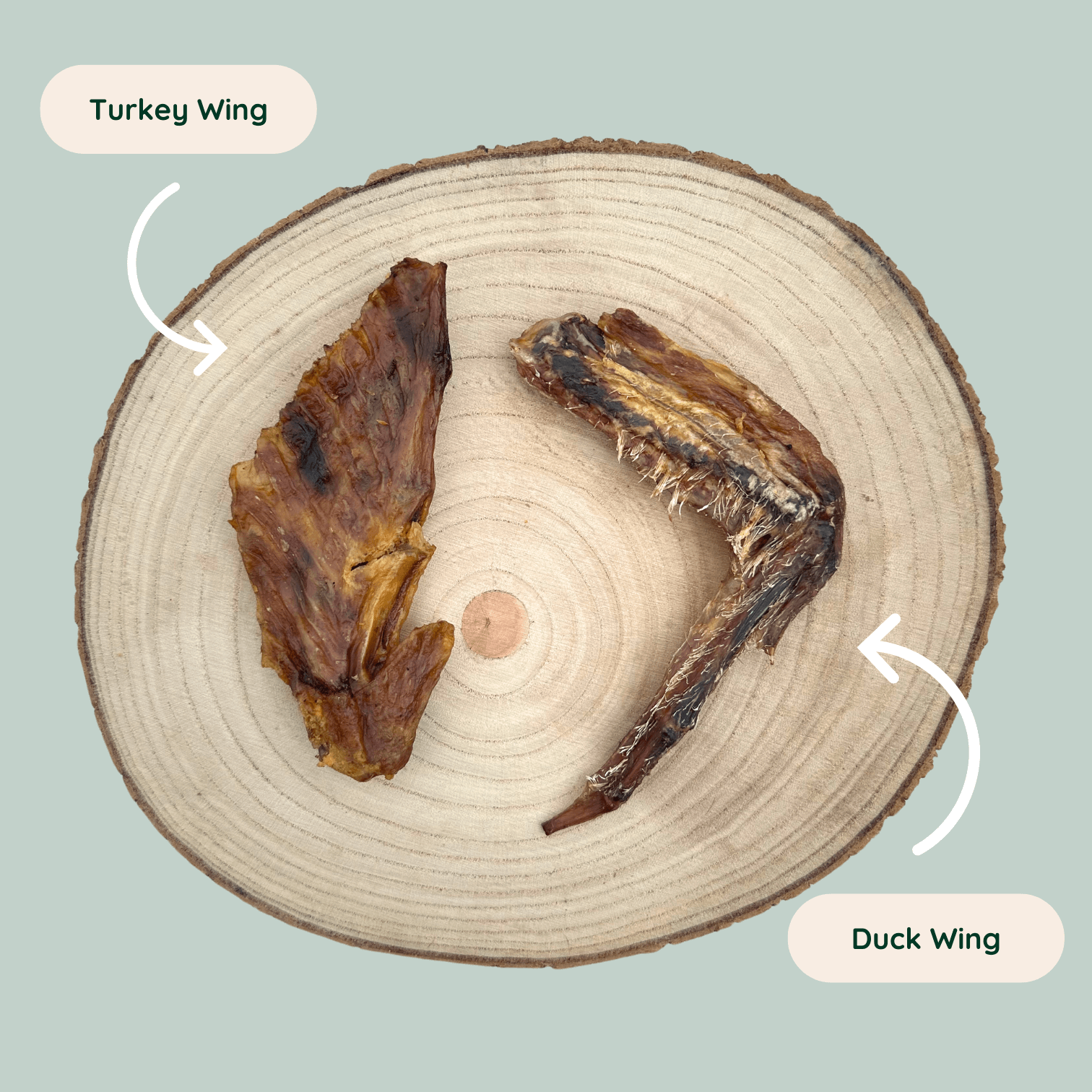 A dried turkey wing and a dried duck wing dog chew on a wooden slice plate against a jade green background.