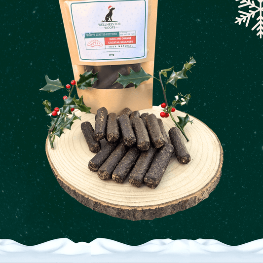 A pile of duck and orange cocktail sausages for dogs in front of a filled pouch of treats and two sprigs of decorative holly. All on a wooden slice plate against a dark green starry sky, with a snowflake in the top right hand corner and a blanket of snow across the bottom.