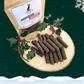 A pile of duck and orange cocktail sausages for dogs in front of a filled pouch of treats and two sprigs of decorative holly. All on a wooden slice plate against a dark green starry sky, with a snowflake in the top right hand corner and a blanket of snow across the bottom.