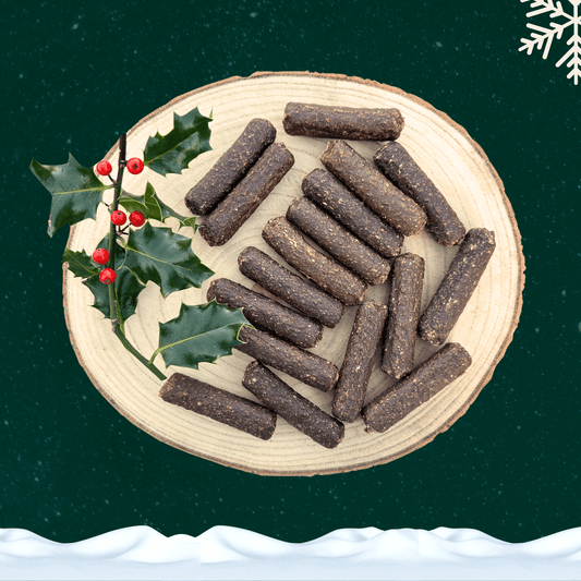 A pile of duck and orange cocktail sausages for dogs next to a sprig of decorative holly. All on a wooden slice plate against a dark green starry sky, with a snowflake in the top right hand corner and a blanket of snow across the bottom.