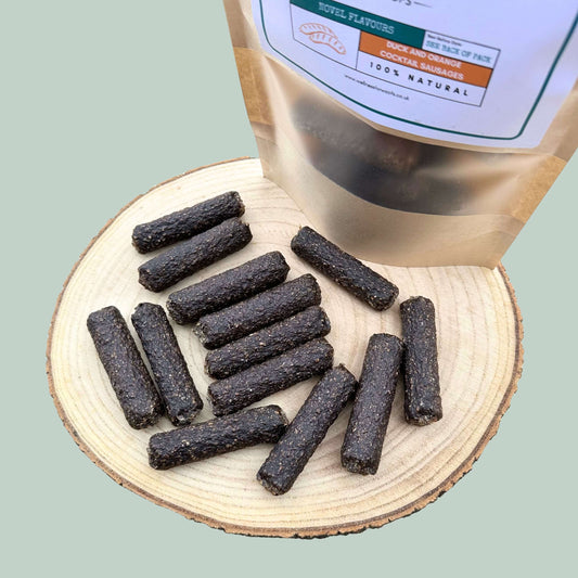 Duck and orange dog treats on a wooden board with a transparent packaging bag labelled '100% Natural' on a light green background.