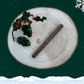 A single guinea fowl sausage stick for dogs lay on a wooden plate next to a piece of holly. The plate sits against a starry green background, a snowflake in the top right corner and snow across the bottom.