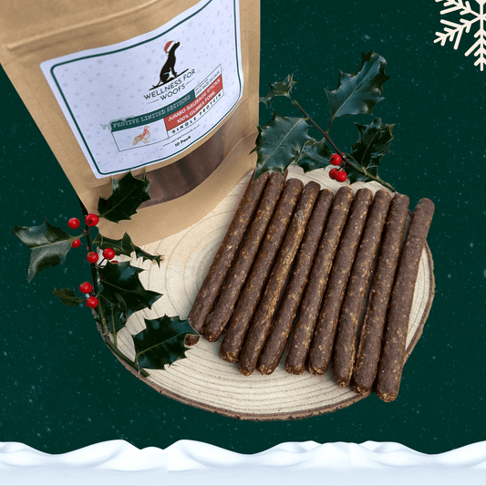 Pack of 10 guinea fowl sausage dog treats next to 10 loose sausages on a wooden plate with 2 pieces of festive holly. All against a green starry background with a snowflake in the top right corner and snow across the bottom.