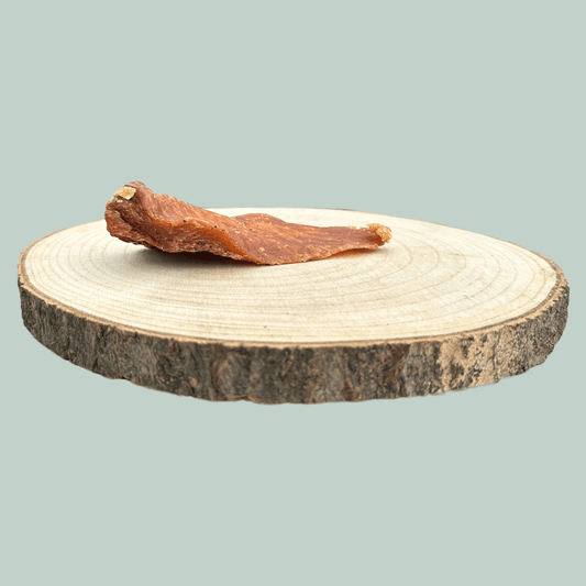 Side view of a single dried chicken fillet natural and healthy dog chews on a wooden slice plate against a jade green background