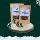 Two pouches of sausage slice dog treats on a wooden plate, standing behind a sprig of holly. All set against a dark green starry sky background with a snowflake in the top right hand corner and a blanket of snow across the bottom.