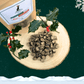 A large pile of 3 bird sausage slices for dogs on a wooden slice plate. Behind the pile stands a pouch full of dog treats and two sprigs of decorative holly. The plate and pouch stand against a dark green starry sky background with a snowflake in the top right hand corner and a blanket of snow across the bottom.