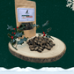 A large pile of 3 bird sausage slices for dogs on a wooden slice plate. Behind the pile stands a pouch full of dog treats and two sprigs of decorative holly. The plate and pouch stand against a dark green starry sky background with a snowflake in the top right hand corner and a blanket of snow across the bottom.