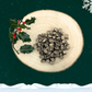 A large pile of 3 bird sausage slices for dogs on a wooden slice plate. Behind the pile stands a sprig of decorative holly. The plate stands against a dark green starry sky background with a snowflake in the top right hand corner and a blanket of snow across the bottom.