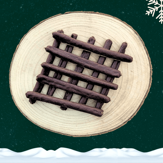 10 venison sausage dog treats stacked neatly on a wooden plate. All against a green starry background with a snowflake in the top right corner and snow across the bottom.