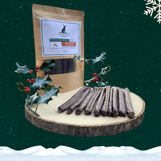 Pack of 10 venison sausage dog treats next to 10 loose sausages on a wooden plate with a piece of festive holly. All against a green starry background with a snowflake in the top right corner and snow across the bottom.