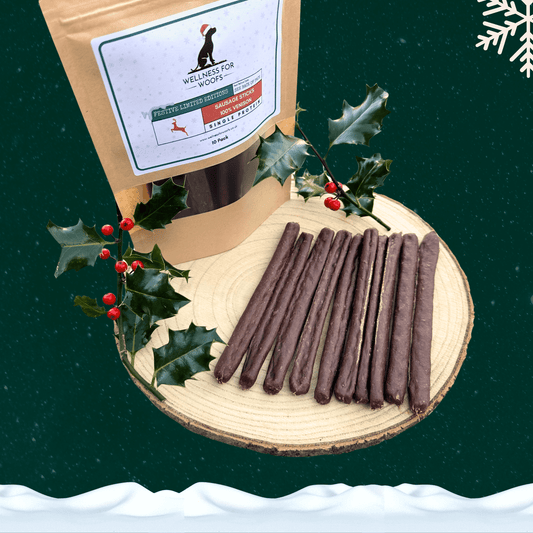 Pack of 10 venison sausage dog treats next to 10 loose sausages on a wooden plate with a piece of festive holly. All against a green starry background with a snowflake in the top right corner and snow across the bottom.