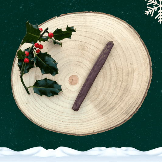 A single venison sausage dog treat on a wooden plate next to a piece of holly. All against a green starry background with a snowflake in the top right corner and snow across the bottom.